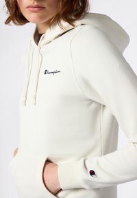 Lichtcrème hoodie gemaakt van zacht materiaal, met een kangoeroezak, verstelbare trekkoord en een klein geborduurd logo op de borst.