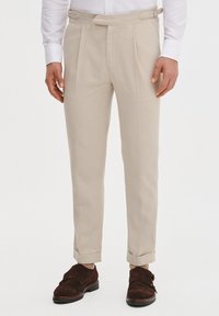 Beżowe spodnie taliowane z płaskim przodem, bocznymi zakładkami i mankietami. Wykonane z lekkiego materiału, charakteryzujące się gładką teksturą i prostymi liniami.