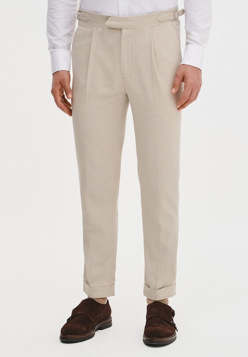 Beżowe spodnie taliowane z płaskim przodem, bocznymi zakładkami i mankietami. Wykonane z lekkiego materiału, charakteryzujące się gładką teksturą i prostymi liniami.