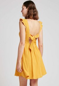 Robe jaune avec des bretelles volantes, dos nu et un détail en nœud. Le tissu a une finition texturée et une jupe évasée et fluide.