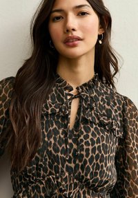 Joven con cabello largo y oscuro que lleva pendientes de aro y una blusa marrón con estampado de leopardo, con nudo frontal y escote con volantes.