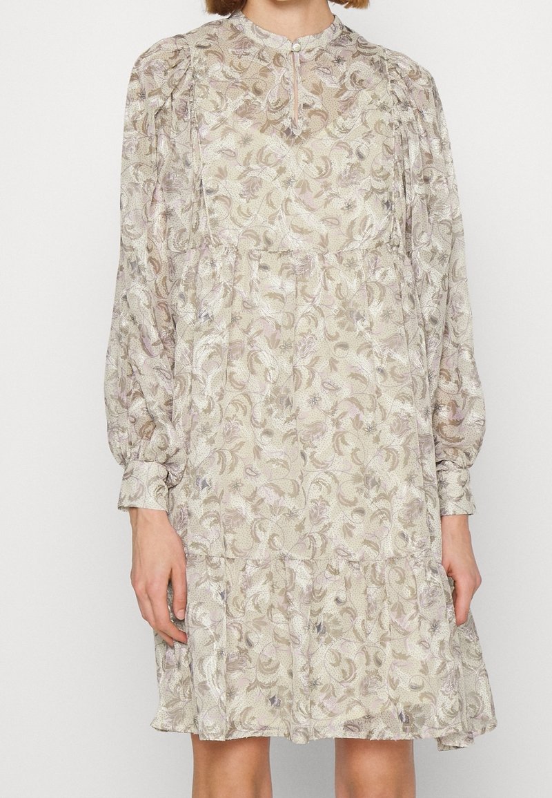 Femme portant une robe légère à manches longues à motifs floraux, avec des épaules bouffantes et un col montant avec une ouverture en forme de trou de serrure fermée par un bouton.