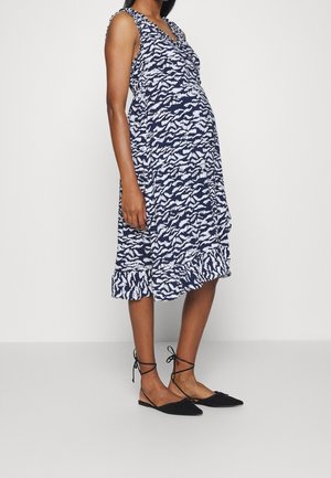 Kobieta w ciąży ubrana w bezrękawową, granatowo-białą sukienkę o długości do kolan w wzory, z czarnymi balerinami sznurowanymi.