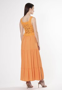 Orange maxi kjole med en struktureret hækle top, lagdelt skirt og glat stof. Bagfra ses åbent lace detalje og en svajende silhuet.