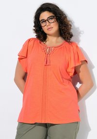 Korálová blůza s krátkým rukávem a šněrovacím výstřihem, doplněná o třásně. Měkká tkanina a uvolněný střih. V kombinaci s olivově zelenými kalhotami.