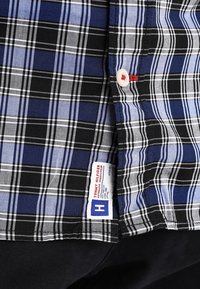 Camicia a quadri blu, nera e bianca con un bottone bianco e un'etichetta Tommy Hilfiger sopra pantaloni scuri.