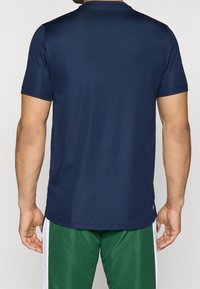 Man i en marinblå kortärmad skjorta och gröna sportshorts med vita ränder, sedd bakifrån mot en enfärgad bakgrund.