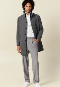 Abrigo de lana gris con cuello alto, cierre de botones y bolsillos laterales, combinado con pantalones gris claro y zapatillas blancas. Textura suave.