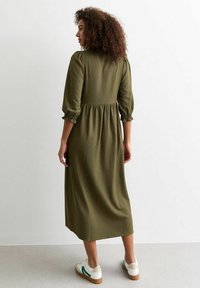 Robe midi vert olive en tissu texturé, avec des manches bouffantes, des poignets élastiques et une taille smockée. Associée à des baskets blanches et des accents verts.