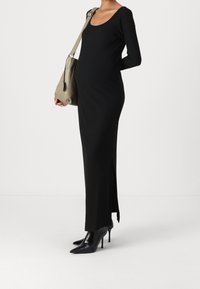 Schwarzes geripptes Maxikleid mit langen Ärmeln, einem runden Ausschnitt und seitlichem Schlitz, kombiniert mit einer grauen Schultertasche und schwarzen Stiletto-Absätzen.