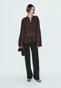 Donna in blusa trasparente marrone scuro con volant, pantaloni neri a gamba larga, che tiene una pochette marrone scuro, in piedi contro uno sfondo bianco semplice.