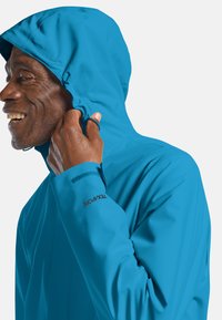 Giacca blu con cappuccio resistente all'acqua, con una texture liscia, cappuccio regolabile e logo nero sulla manica. Presenta una vestibilità aderente e maniche lunghe.