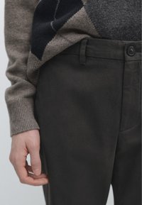 Personne portant un pull à losanges marron et bleu marine avec un pantalon noir et une taille boutonnée, main détendue sur le côté.