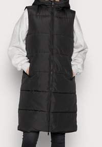 Person som bär en lång svart pufferväst med huva, grå sweatshirtärmar synliga, händerna i västfickorna och svarta byxor.