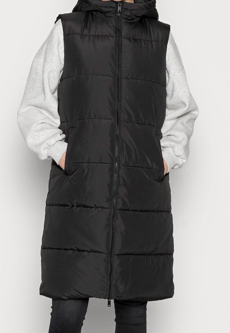 Person som bär en lång svart pufferväst med huva, grå sweatshirtärmar synliga, händerna i västfickorna och svarta byxor.