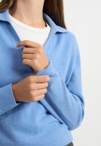 Jasnoniebieski, miękki sweter z dekoltem w serek i kołnierzykiem. Posiada prążkowane mankiety oraz luźny krój na białej koszuli pod spód.