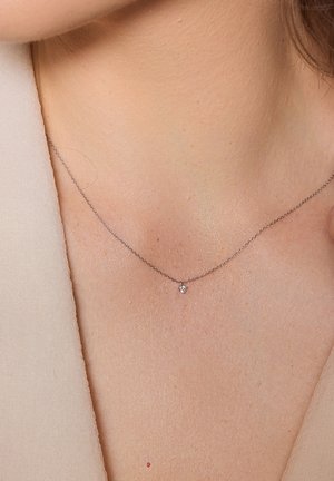 Srebrny łańcuszek z małym diamentowym wisiorem leżący na skórze. Delikatny wzór, połyskujące wykończenie, krótka długość, z drobnymi detalami łańcuszka.