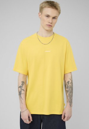 Jeune homme portant un T-shirt jaune surdimensionné avec le texte "CAPITAINE", un collier en chaîne argentée et des tatouages sur les deux avant-bras, sur un fond gris.