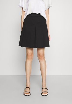 Mujer con falda negra plisada hasta la rodilla, blusa blanca de manga corta y sandalias negras de punta abierta, sobre un fondo liso.