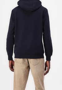 Felpa blu navy in misto cotone, con tasca a marsupio, polsini e vita ribattuti. Indossata con pantaloni beige chiaro e texture.