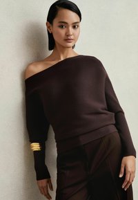 Dunkelbrauner, schulterfreier, gerippter Pullover mit langen Ärmeln, der an einem Ärmel mit einem goldenen Akzent versehen ist. Kombiniert mit dunklen, glatten Hosen.