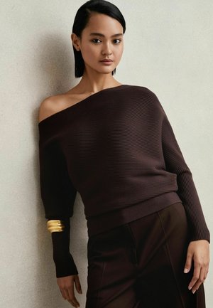 Dark brown off-the-shoulder ribbed sweater with long sleeves, featuring a gold accent on one sleeve. Paired with dark, smooth trousers.
