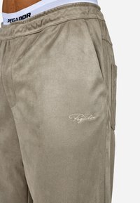 Beige suede joggers with an elastic waistband and embroidered "Pegador" logo. Features side pockets and smooth, soft texture.