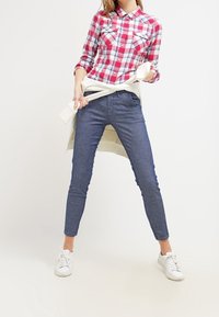 Rutig röd och vit flanellskjorta med två bröstfickor, kombinerad med blå slim fit-jeans och vita sneakers. Vit tröja knuten i midjan.