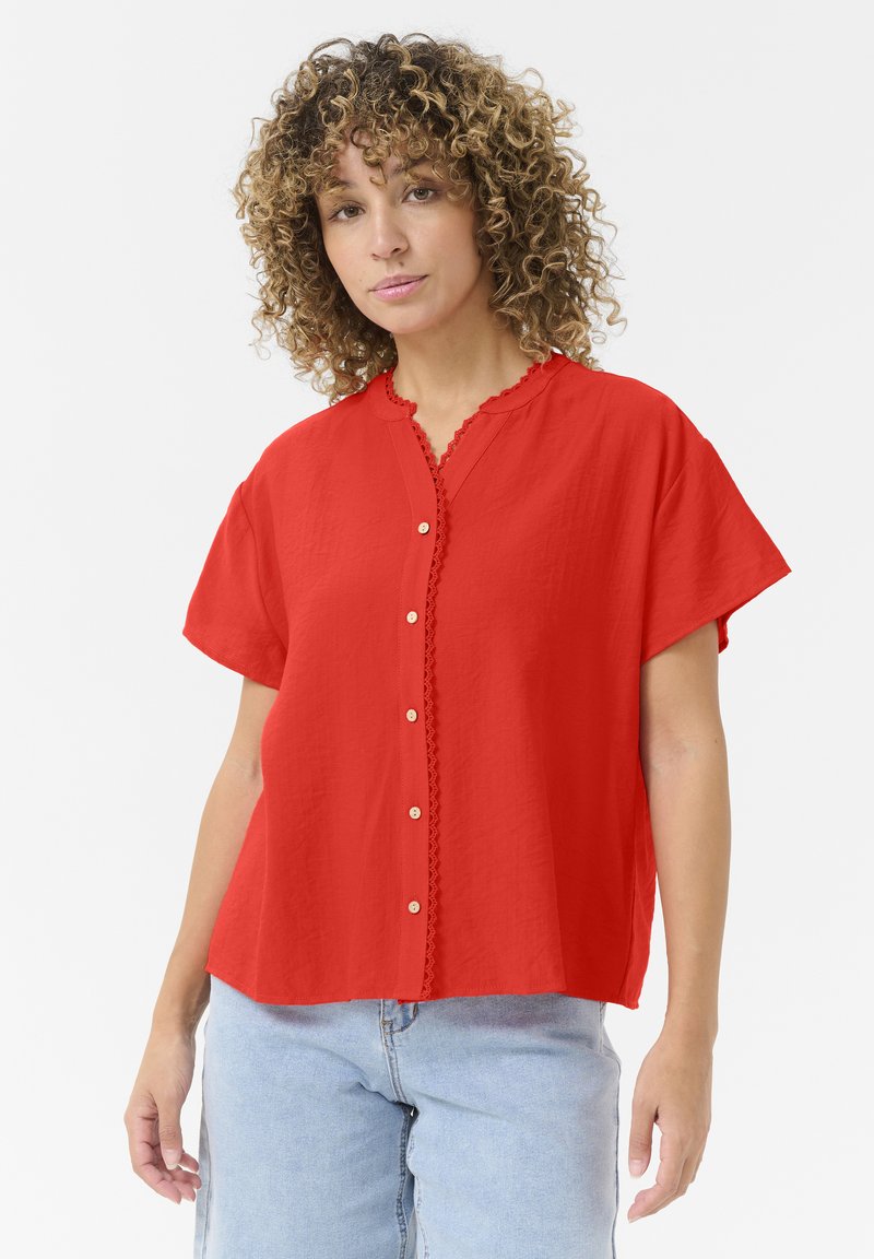 Rood shirt met korte mouwen, voorzien van een gegolfde kraag, knoopsluiting aan de voorkant en een textuurstof. Heeft decoratieve stiksels langs de halslijn.
