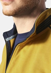 Gelbe Jacke mit hohem Kragen und marineblauen Akzenten, mit einem Reißverschluss und kontrastierenden Nähten. Nahaufnahme des Kragenbereichs.