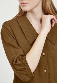 Chemise marron à boutons avec un grand col, en tissu texturé et aux manches froncées. Comprend des boutons de couleur claire et une coupe décontractée.