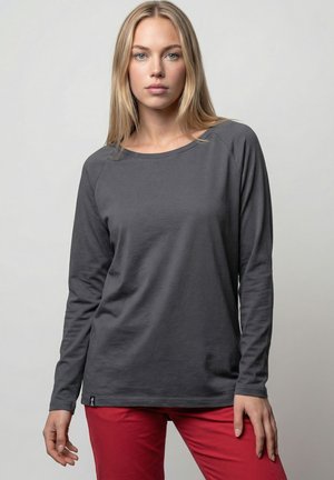 Blonde woman wearing a dark gray long-sleeve shirt and red pants, standing against a plain light gray background.