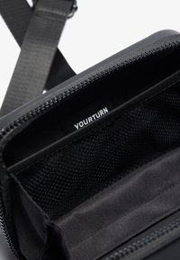 Sort stofpose med lynlåslukning, der har en mesh-lomme indeni og en label med teksten "YOURTURN". Glat tekstur, kompakt rektangulær form.