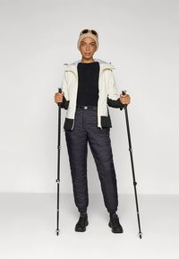 Utendørs antrekk med hvit og svart jakke, svart skjorte og polstrede svarte bukser. Tilbehør inkluderer vandrestaver og solbriller.