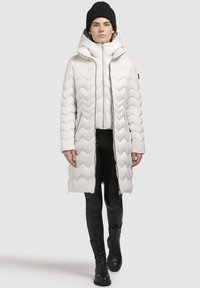 Vit pufferjacka med chevronmönster, hög krage, dragkedja och avtagbar huva, kombinerad med svarta leggings och stövlar.