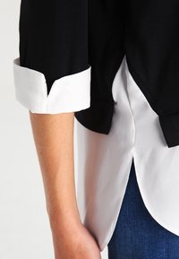 Svart och vit blus med trekvartslånga ärmar, i ett mjukt bomullsmaterial. Har en vit skjort-svans fåll och kontrasterande manschettdetaljer.