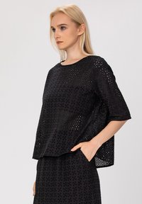 Haut noir à manches courtes en tissu texturé avec des motifs découpés. Coupe légèrement ample avec un col rond.