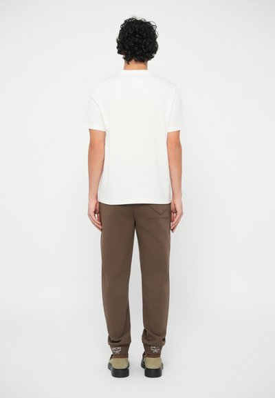 Άνδρας με σγουρά μαλλιά στέκεται με την πλάτη στραμμένη, φορώντας ένα λευκό κοντομάνικο πουκάμισο, καφέ παντελόνια και μπεζ με μαύρα παπούτσια.