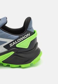Chaussure de course pour homme avec une tige en mesh gris, un contrefort noir au talon et une semelle extérieure verte. Comprend un design texturé et une marque sur le côté.
