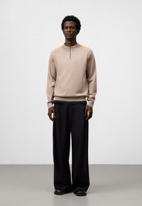 Maglione beige con colletto alto e dettaglio con zip, abbinato a pantaloni neri larghi e scarpe nere. Tessuto liscio e vestibilità comoda.