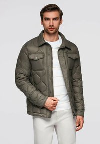 Uomo che indossa una giacca trapuntata verde sopra un maglione bianco e pantaloni bianchi, in piedi contro uno sfondo grigio chiaro uniforme.
