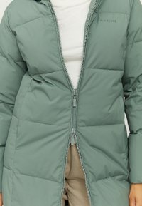 Padded coat in muted green, featuring a full zipper, a hood, and quilted texture. Logo "MAZINE" on left shoulder.