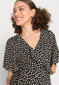 Vestido preto com padrão de folhas brancas e amarelas, decote em V, mangas curtas e detalhes franzidos sutis na gola. Cabelo encaracolado, brincos de argola.