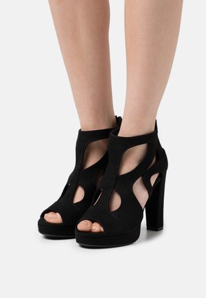 Sandales à talons hauts en daim noir avec design à découpes, bouts ouverts et semelle plateforme. Elles comportent un talon épais et carré ainsi qu'une fermeture éclair latérale.