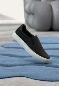 Baskets noires à enfiler avec semelle blanche, posées au-dessus d'un tapis bleu texturé et d'un canapé gris capitonné dans un salon moderne.