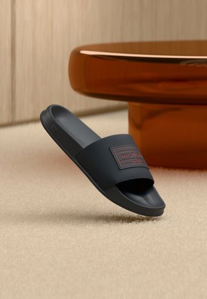 Schwarzer Slide-Sandal mit texturiertem Riemen und rotem „Hilfiger“-Logo, schwebt über einem beigen Teppich in der Nähe eines runden braunen Tisches.