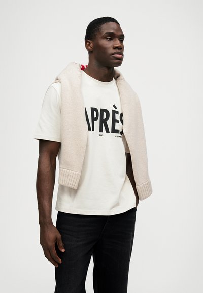 T-shirt creme com "APRÈS" em texto preto em negrito, combinada com um suéter de malha cinza claro colocado sobre os ombros, e jeans escuros.