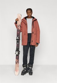 Μπουφάν για σκι σε χρώμα σκουριάς με μαύρο εσωτερικό, συνδυασμένο με μαύρο παντελόνι χιονιού και μπότες για σκι· κρατώντας ένα ανοιχτό καφέ πέδιλο σκι με μαύρα δέστρες.