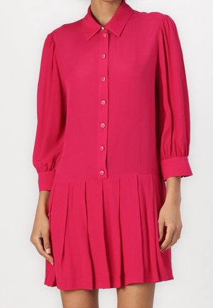 Femme portant une robe boutonnée rose vif avec de longues manches bouffantes et une jupe plissée sur un fond uni.