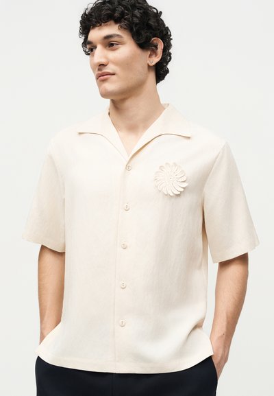 Άνδρας που φοράει λευκοκρεμ κοντομάνικο πουκάμισο με κουμπιά και ένα λουλουδάτο δαντελένιο καρφίτσα στο αριστερό στήθος, στέκεται με τα χέρια στις τσέπες.